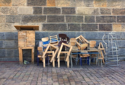 The image shows an assortment of wooden chairs and tables arranged in front of a textured brick wall, likely outdoors on a paved surface. The chairs vary in design, with some featuring light wood finishes and others darker or painted surfaces. Several chairs are stacked or leaned against each other, creating a cluttered appearance. To the left, there is a tall wooden shelving unit or cabinet with an open top, and a small wood crate or box is placed on top. On the far right, there is a metal wire chair or stool with a round seat, standing slightly apart from the other furniture. The scene reflects a collection of used or discarded outdoor furniture, possibly awaiting removal or disposal, consistent with rubbish removal services provided by House Clearance Epping in the local Epping area. The overall atmosphere suggests an outdoor storage or clearing situation, with natural daylight illuminating the scene and highlighting the varied textures of the wood and brickwork.
