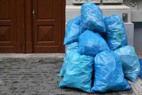 A collection of multiple large blue plastic rubbish bags filled with waste, stacked on a paved driveway in front of a brown wooden door. The bags are loosely tied at the top, with some showing slight bulges indicating the contents inside. The driveway surface consists of grey paving stones, and the bags are positioned along the edge of a residential property, suggesting a rubbish removal service taking place in Epping. The background includes partial views of a white wall and an outdoor electrical socket while a grey utility box is visible on the right side. The scene appears to be outdoors during daytime with natural light illuminating the area. This image reflects the typical rubbish collection or house clearance activities carried out by House Clearance Epping, designed to assist with the disposal of household waste efficiently, particularly in the local postcode area.