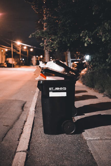 A black wheeled rubbish bin positioned on a residential pavement at night, with the label 'St. John's' visible on the front. The bin is filled with various discarded items, including cardboard boxes,