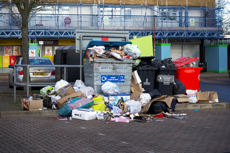 A black wheeled rubbish bin positioned on a residential pavement at night, with the label 'St. John's' visible on the front. The bin is filled with various discarded items, including cardboard boxes,