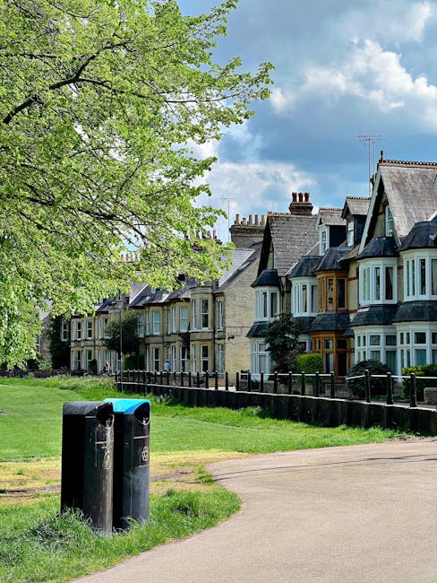 In a peaceful residential area, two black public bins for rubbish disposal are positioned on a grassy verge beside a paved footpath. The bins are cylindrical with rounded tops, one slightly taller tha