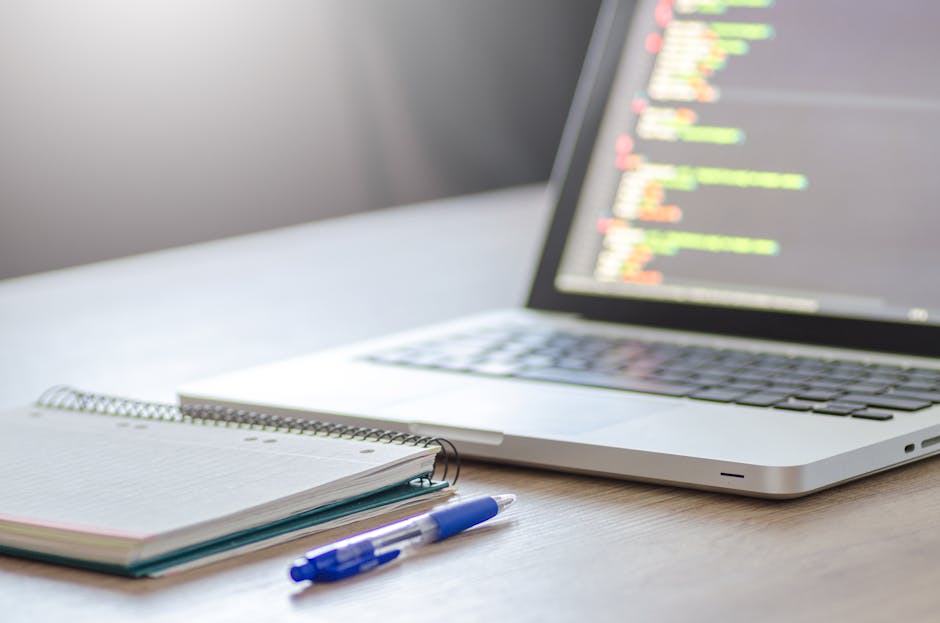 A close-up view of a silver laptop placed on a light wooden desk, with its screen displaying lines of programming code in various colors, including yellow, white, and blue. To the left of the laptop, 