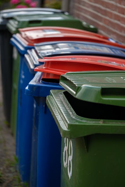 A waste collection worker wearing a high-visibility yellow and red uniform is standing at the rear of a large red rubbish truck, which is parked on a paved road. The truck features a hydraulic compact